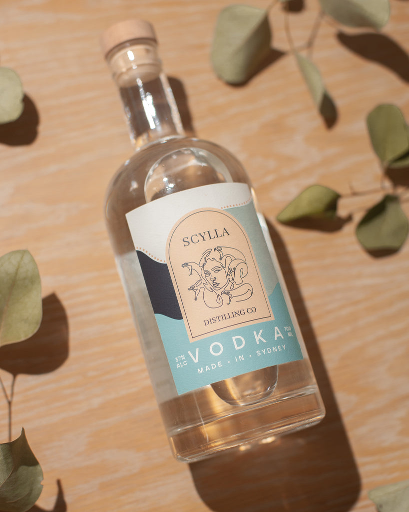 Scylla Vodka bottle laying on a wood finish surface with dried leaves on either side.