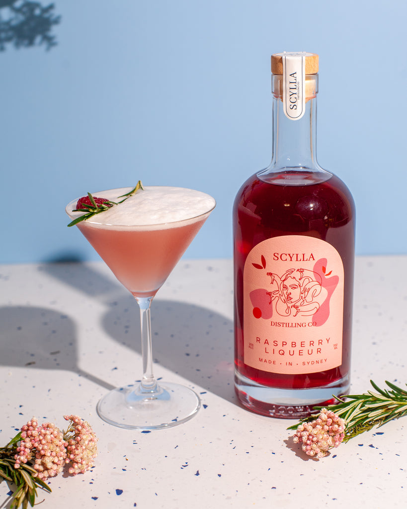 Scylla Raspberry liqueur is shown on the right and on the left is a French Martini with a sprig of rosemary and a raspberry as garnish. The background is a pale blue.